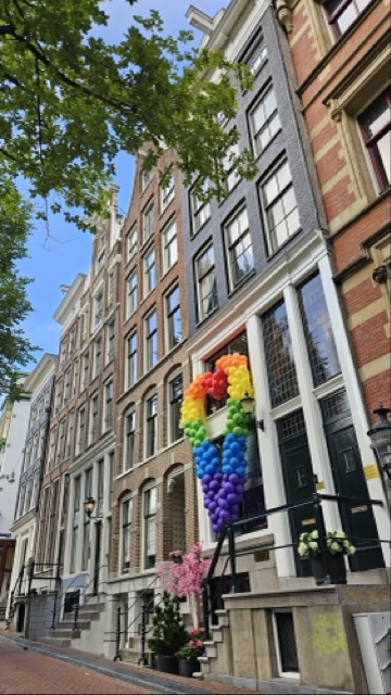 Building decorated with a heart-shaped rainbow balloon display during Amsterdam Pride, with people celebrating LGBTQ+ pride in the street.