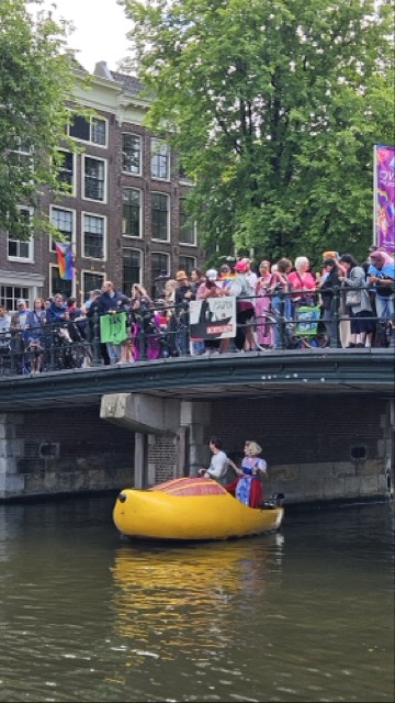 The opening boat of Amsterdam Pride, shaped like a giant wooden clog shoe with a "Holland Antje" figure, floats on a canal, surrounded by celebrating people with rainbow flags.