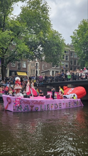Boat decorated in pink sailing in the Amsterdam Canal Pride parade, with people celebrating LGBTQ+ pride along the canals.