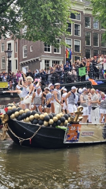 A boat filled with celebrating people and rainbow flags floats on an Amsterdam canal during Pride and audience in the background.