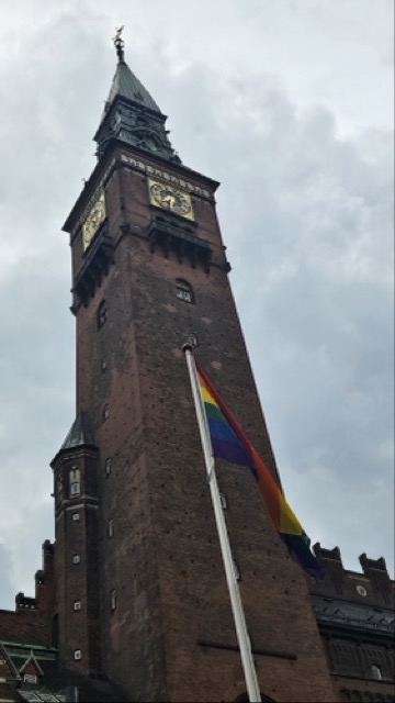 A photograph of the Copenhagen City Hall building (Rådhuspladsen) with a large rainbow flag flying in front of it.