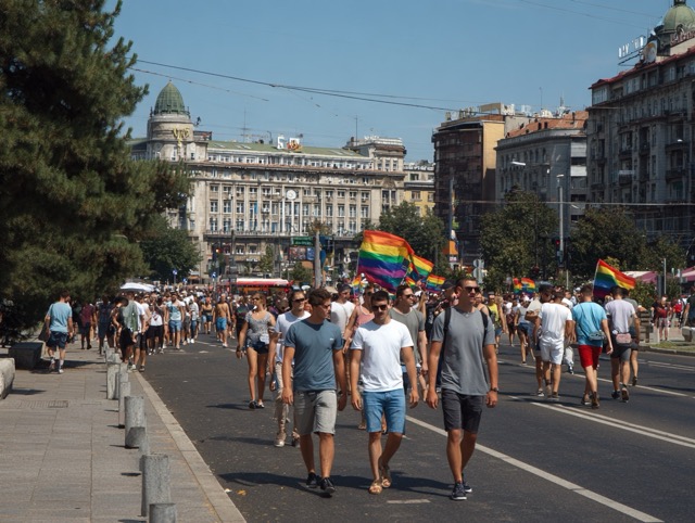 People gathered for an LGBT pride event in Belgrade, Serbia, with participants holding rainbow flags.