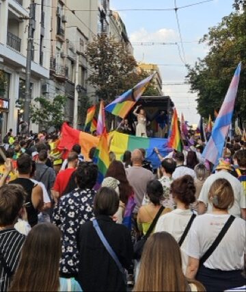 Crowd of people walking in the streets of Belgrade during the annual Pride parade, holding rainbow and trans flags.