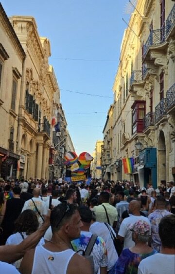 Crowds with rainbow flags celebrating Malta Pride parade against a backdrop of historic Maltese architecture.