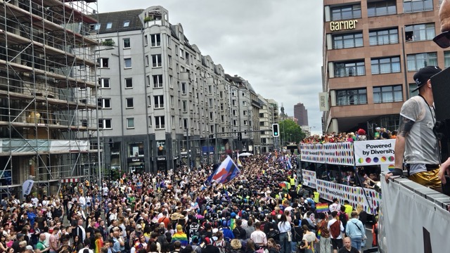 Colorful floats and crowds celebrating Berlin Pride parade, with people waving rainbow flags and dancing through the city streets.