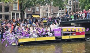 Colorful boats and crowds celebrating Amsterdam Canal Pride parade, with people waving rainbow flags along the canals.