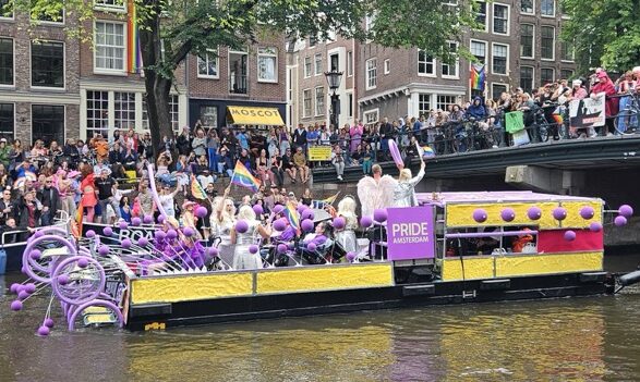 Colorful boats and crowds celebrating Amsterdam Canal Pride parade, with people waving rainbow flags along the canals.