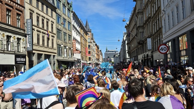 Colorful floats and crowds celebrating Copenhagen Pride parade, with people waving rainbow flags and dancing through the city streets.