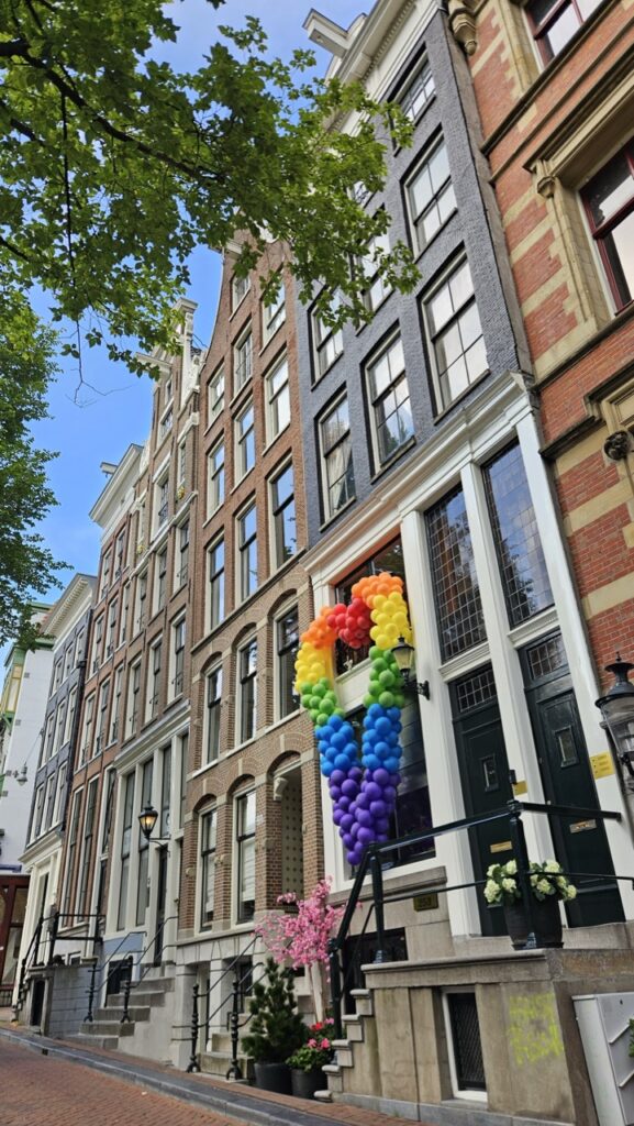 Building decorated with a heart-shaped rainbow balloon display during Amsterdam Pride, with people celebrating LGBTQ+ pride in the street.