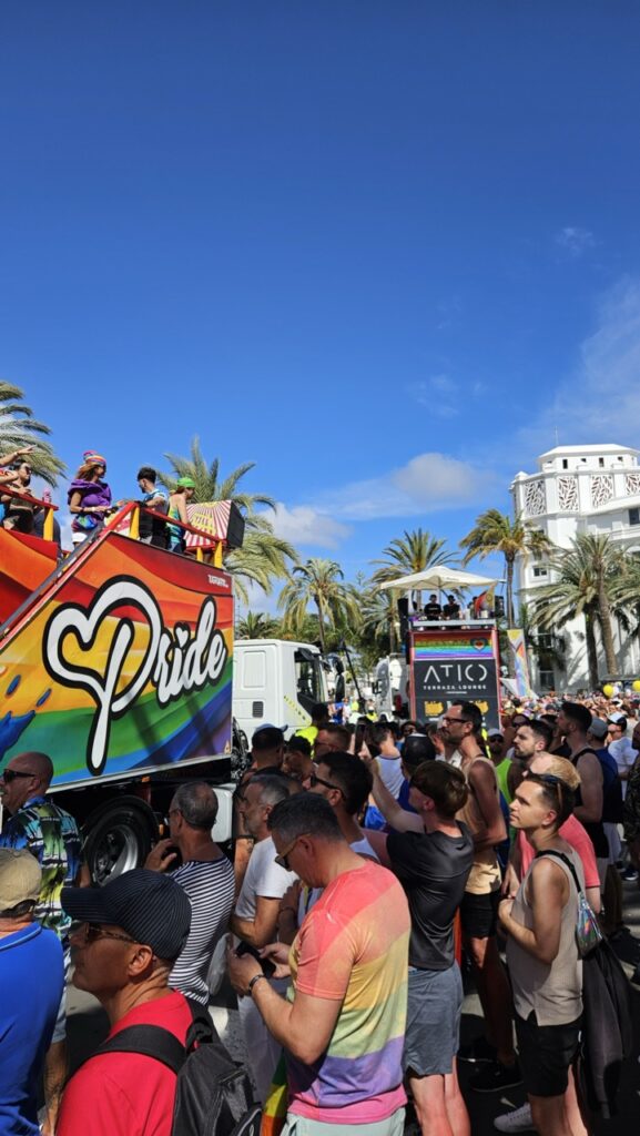 People celebrating at Maspalomas Pride in Gran Canaria, with rainbow flags and a festive crowd enjoying the sunny outdoor event.
