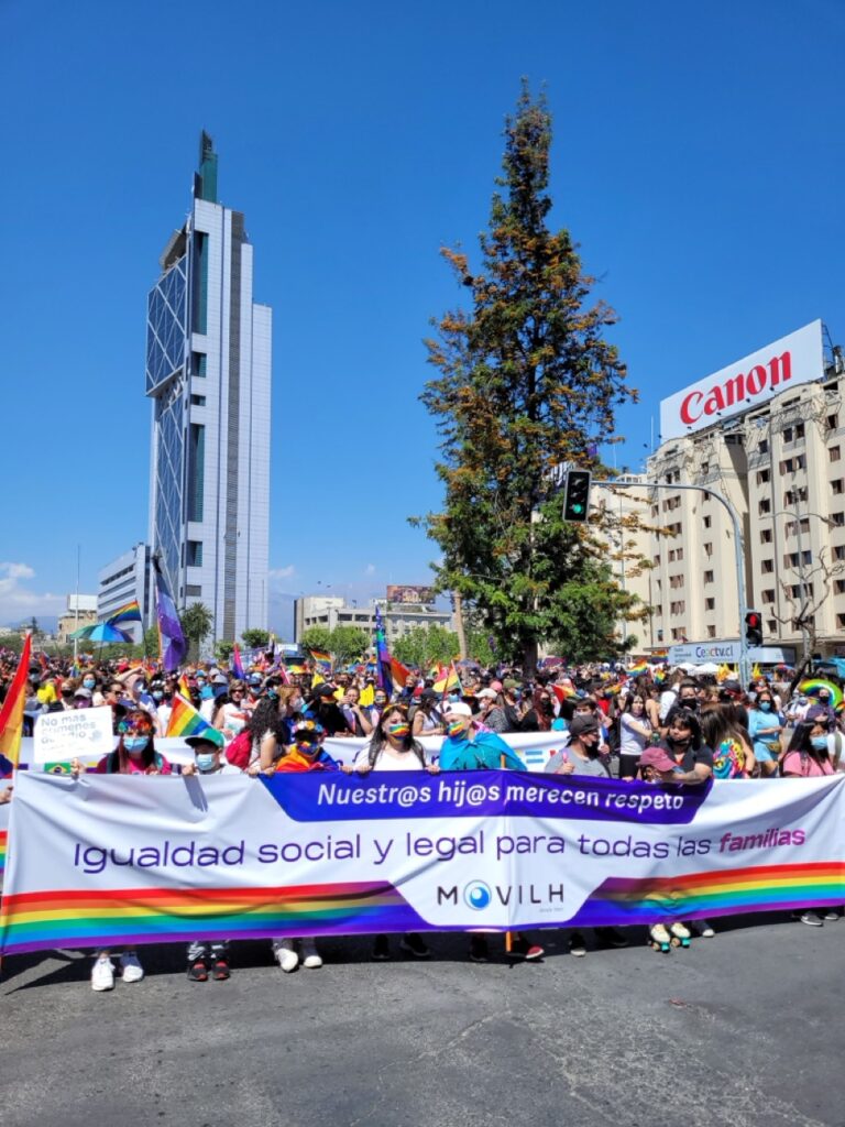 People marching in a South American Pride parade holding a banner, with crowds celebrating LGBTQ+ pride in the streets.