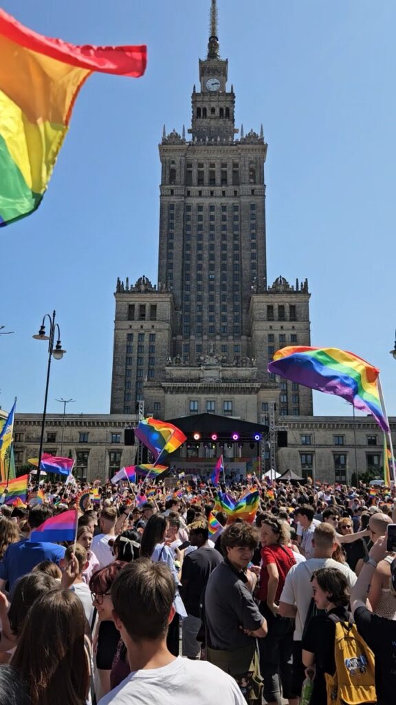 Crowd at Warsaw Pride village stage concert with the Palace of Culture and Science in the background, celebrating LGBTQ+ pride.
