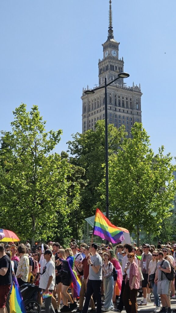 People marching during Warsaw Pride with rainbow flags, with the Palace of Culture and Science visible in the background.