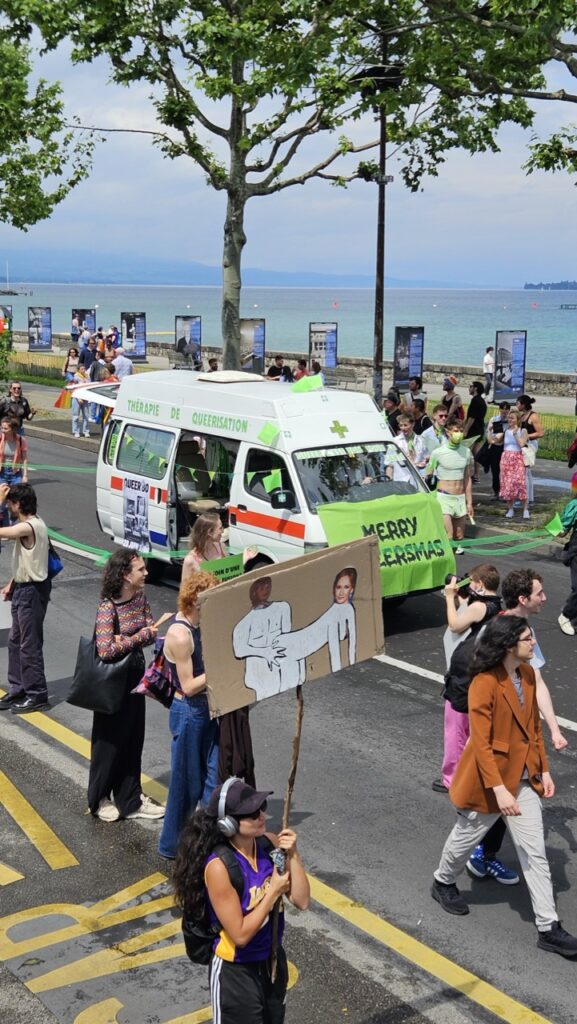 People holding political posters during Geneva Pride with Lake Geneva in the background, celebrating LGBTQ+ rights.