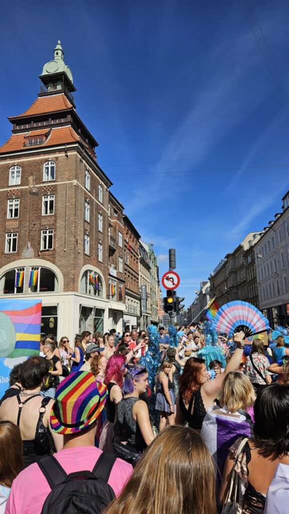 Crowd marching through Copenhagen city centre during Copenhagen Pride, with people celebrating LGBTQ+ pride and waving rainbow flags.