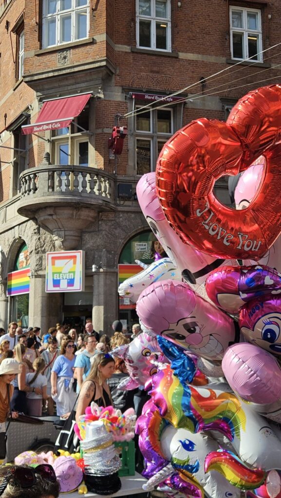 Crowd celebrating Copenhagen Pride in the city centre, with a convenience store decorated in rainbow colors.