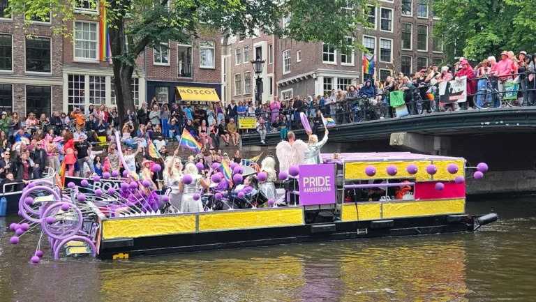 Colorful boats and crowds celebrating Amsterdam Canal Pride parade, with people waving rainbow flags along the canals.