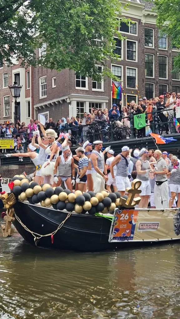 A boat filled with celebrating people and rainbow flags floats on an Amsterdam canal during Pride and audience in the background.