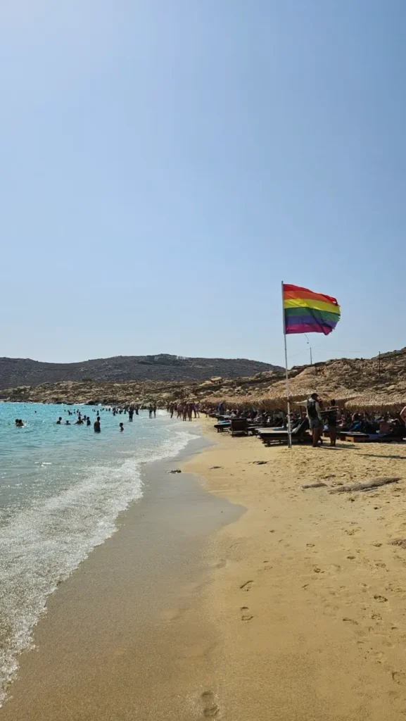 People celebrating Mykonos Pride at a popular gay beach with rainbow flags and the Aegean Sea.