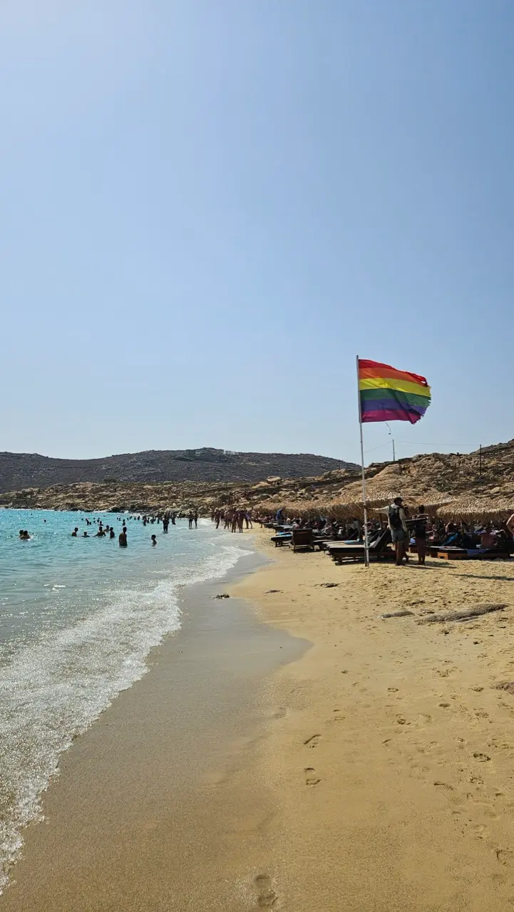 People celebrating Mykonos Pride at a popular gay beach with rainbow flags and the Aegean Sea.